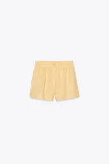 Short de tiro medio de lino 100%, color amarillo claro. Presenta cintura elástica, cierre frontal con botones y bolsillos laterales ocultos en costura y de plastrón en la espalda. El bajo tiene aberturas laterales.