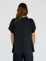 Camisa negra de manga corta con cuello redondo y detalle de volados frontales. Presenta cierre central con botones de madera y terminaciones con puntilla en el escote.