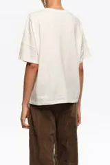 Remera de manga corta color crudo, con cuello redondo y diseño holgado. Presenta un bordado en el pecho con motivo verde y amarillo y la inscripción de la marca.