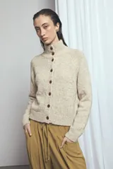 Cardigan de punto color beige melange, de cuello alto y cierre frontal con botones marrones. Cuenta con mangas largas y diseño clásico.