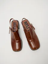 Zuecos de cuero vacuno con acabado en charol, color beige. Presentan un diseño clásico con taco cuadrado de madera de 5 cm y correa trasera ajustable con hebilla metálica.