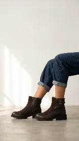 Bota urbana de caña media, confeccionada en una combinación de cuero gastado y gamuza. Presenta una solapa frontal en gamuza, cierre lateral y una tira decorativa con hebilla metálica en el tobillo. Cuenta con una suela de goma robusta y dentada.