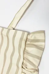 Bolso tipo tote bag de tela con diseño de rayas verticales en tonos beige y gris, decorado con volados en los laterales y la base. Incluye dos asas largas para llevar al hombro y una etiqueta con logo en el frente.