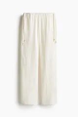 Pantalón de corte holgado y piernas amplias, confeccionado en tela con textura arrugada. Presenta tiro bajo con cintura elástica y cordones laterales ajustables con cuentas decorativas en los extremos.
