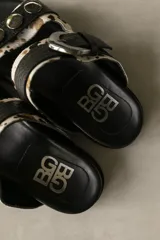 Sandalias de cuero de doble tira, con estampa animal print tipo vaca en tonos blanco, negro y marrón, adornadas con tachas metálicas en la tira superior y una hebilla grande en la tira inferior.