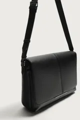 Bandolera de diseño rectangular en color negro con acabado texturizado. Cuenta con una solapa frontal con detalle metálico, cierre imantado y asa de hombro ajustable. El interior incluye un bolsillo plano y otro con cierre, además de un bolsillo plano adicional en la parte frontal.