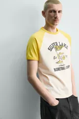 Camiseta de corte relajado con mangas raglán cortas en color amarillo y cuerpo en color crema. Presenta un estampado gráfico frontal con temática de campamento de básquetbol y un cocodrilo.