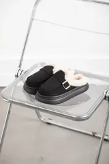 Zueco de gamuza color negro con interior forrado en corderito sintético, suela de goma tipo plataforma y detalle de hebilla metálica dorada en el empeine.