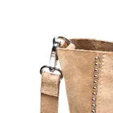 Cartera tipo tote bag de gamuza en color tostado, con doble asa de mano y correa ajustable extraíble. Presenta detalles de tachas metálicas en los laterales y un logo circular grabado en el centro.