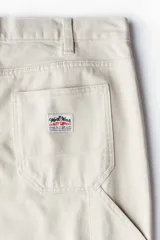 Pantalón worker de sarga de algodón gruesa color beige, con piernas rectas y corte relajado. Presenta cintura clásica con cierre de botón, bolsillos al bies, bolsillo monedero, bolsillos traseros, trabilla funcional en un lateral y bolsillo cargo dividido en dos en el otro.
