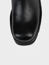 Bota negra de cuero hasta media pantorrilla, con plataforma y taco metálico plateado. Presenta detalles de tachas plateadas y correa con hebilla metálica.