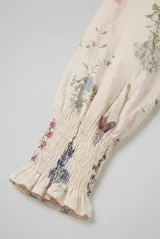 Camisa de manga larga color arena con delicado estampado botánico de flores silvestres. Presenta cuello alto con volados, escote en V con cierre frontal de botones y puños ajustados con nido de abeja.