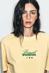 Camiseta amarilla de corte oversize, con cuello redondo, manga corta y estampa frontal en color verde con texto alusivo a Brasil Ipanema y pequeños símbolos.