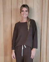 Remerón de textura acanalada en color gris claro, con cuello redondo, mangas largas y bolsillo frontal en el pecho. Presenta un diseño holgado con corte asimétrico, siendo más largo en la parte posterior.