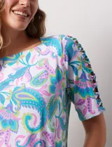 Remera de algodón estampado con diseño paisley en tonos celeste, rosa y amarillo. Tiene cuello redondo y mangas cortas con detalle de entrelazado y aberturas en los hombros.
