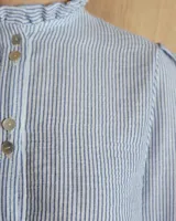 Camisa de manga tres cuartos color beige con cuello con volados y botones frontales. Presenta un detalle de color negro en la parte superior de las mangas.