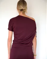 Vestido corto color bordó, con diseño de escote asimétrico y detalle de fruncido lateral con pequeñas argollas metálicas en uno de los hombros. Presenta un corte ajustado en la parte inferior y una caída holgada en el torso.