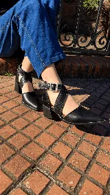 Zapatos negros de cuero con punta fina, taco ancho y diseño destalonado con tiras cruzadas texturizadas y hebilla metálica.