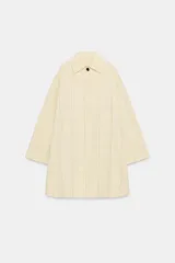 Trench de algodón color amarillo claro con patrón a cuadros sutiles de líneas oscuras. Tiene cuello con solapa, mangas largas, bolsillos delanteros y cierre frontal con botones ocultos bajo solapa.