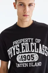 Remera negra de algodón con cuello redondo, manga corta y hombros caídos, de corte holgado. Presenta un estampado gráfico frontal estilo universitario en color blanco con efecto desgastado que lee "PROPERTY OF PHYS. ED. CLASS 1905 STATEN ISLAND".