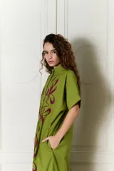 Vestido largo verde militar de popelina de algodón, con diseño oversized, cuello mao, mangas cortas, bolsillos laterales y cierre frontal con botones. Presenta un estampado de flores grandes en la parte superior y franjas horizontales de colores en el ruedo.