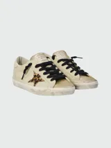 Championes Golden Goose modelo Super Star, con diseño de efecto desgastado. Presentan cuerpo en cuero color crema, puntera y lengüeta con acabado de glitter dorado, estrella lateral con estampado animal print y cordones negros.
