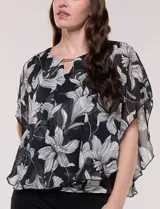 Blusa de gasa semitransparente con hilos de lúrex, diseño estampado floral en tonos blanco, gris y negro, silueta holgada tipo capa y escote redondo con detalle metálico.