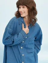 Camisa de denim azul de corte oversize, con mangas largas, cuello clásico, un bolsillo aplicado en el pecho y cierre frontal con botones metálicos.