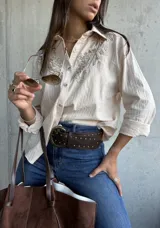 Camisa de color beige con textura rayada vertical, cuello clásico y mangas largas. Presenta un delicado bordado floral en tono sobre tono en la parte superior delantera.