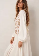 Vestido largo beige con escote en V y mangas largas abullonadas, adornado con delicados bordados florales en tono tostado en la parte frontal y cenefas decorativas a lo largo de la abertura central.