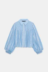 Blusa corta de color celeste con cuello solapa y manga larga abullonada. Presenta un diseño con bordados perforados en blanco en toda la prenda y cierre frontal con botones.