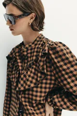 Blusa de manga larga con estampado de cuadros vichy en tonos marrón y negro. Presenta cuello redondo, cierre frontal con botones y detalle de volantes en el pecho.