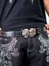Cinto negro de poliuretano con tachas metálicas en los bordes y hebilla grande de metal plateado con diseño de calaveras dobles atravesadas por dagas.
