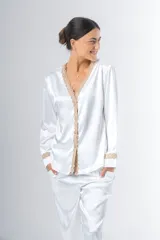 Pijama blanco de satén elastizado, compuesto por una camisa de manga larga con escote en V, abotonada adelante y con puntilla en escote y puños, y un pantalón recto con bolsillos laterales, elástico en la cintura y puntilla en el ruedo.
