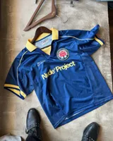 Remera de fútbol estilo vintage de la marca Nude Project, en color azul marino con cuello polo y detalles en amarillo. Presenta inscripciones y un escudo bordado en el pecho.