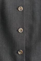 Chaleco largo de vestir color gris oscuro con finas rayas verticales, cuello en V y cierre frontal de botones. Presenta bolsillos delanteros ribeteados y abertura en la parte trasera inferior.