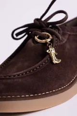 Zapato tipo mocasín acordonado de gamuza color marrón oscuro, con suela de goma color caramelo y detalle de dije metálico dorado en forma de bota colgando de los cordones.