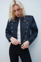 Chaqueta de denim de corte corto, color azul oscuro, con cuello redondo, hombreras y cierre frontal con cremallera. Presenta diseño de inspiración militar con doble botonadura metálica decorativa en el frente, trabillas con botón en los hombros y bajo elástico.
