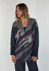 Saco de lana color verde militar con estampado abstracto en tonos verde oliva y detalles bordados en rojo. Tiene cuello solapa, cierre frontal con botones de madera y puños acanalados.