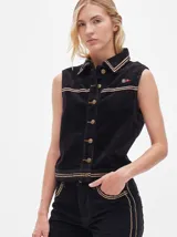 Chaleco de pana negro con cierre frontal de botones metálicos dorados. Presenta detalles de costuras en contraste en el cuello, cartera y canesú, e incluye un pequeño parche con forma de bandera en el pecho.