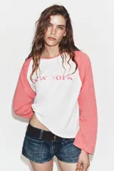 Camiseta de manga larga tipo ranglán con cuerpo blanco y mangas en color rosado. Presenta un estampado frontal con la inscripción 'New York' en tono rosado.