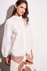 Blusa blanca de algodón con cuello mao, mangas largas abullonadas y detalles de encaje en el canesú, mangas y ruedo. Cierre frontal con botones.