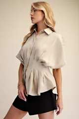 Camisa de popelina color beige con diseño estructurado, presenta pliegues verticales en la cintura que crean un efecto entallado y mangas cortas con volumen. Cuenta con cuello clásico y cierre frontal de botones.