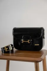 Cartera negra de cuero charolado con solapa y cierre de broche. Presenta un detalle metálico dorado en forma de freno de caballo en la parte frontal y una correa ajustable.