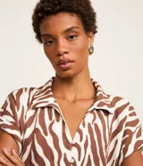 Blusa de manga corta confeccionada en viscosa, con cuello camisero, cierre frontal con botones y detalle de lazo para anudar en el bajo. Presenta un estampado híbrido entre follaje y animal print de cebra en tonos marrón y blanco.