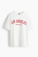 Remera oversized de punto de algodón en color blanco, con cuello redondo, hombros caídos y diseño estampado en el frente con la inscripción "Los Angeles California U.S.A." en color rojo.