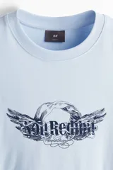 Remera de algodón color celeste con cuello redondo acanalado, mangas cortas y hombros caídos. Presenta un estampado gráfico central con alas y la frase 'No Regret'.