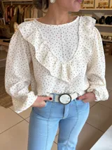 Blusa de tela liviana color crema con estampado floral pequeño en tonos rojizos. Presenta un diseño con volados en el frente y hombros, mangas largas abullonadas con puños elásticos y cuello redondo.