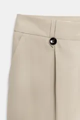 Pantalón de vestir color beige claro, tiro alto, corte recto y ancho. Presenta pinzas frontales con botón negro decorativo y pliegues marcados en la pierna.