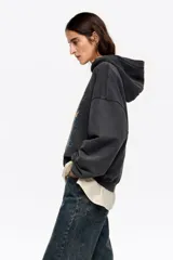 Sudadera con capucha de algodón color negro desgastado, corte globo y manga larga. Presenta un estampado frontal que muestra una escena urbana con edificios, flores amarillas y una bota de lluvia verde sobre un muro de ladrillos.